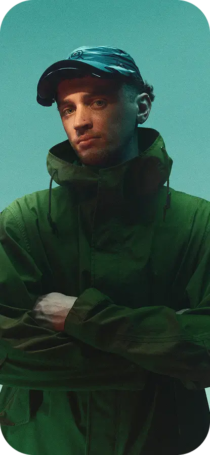 Person wearing a dark green jacket and the Captured Cap, standing against a muted green background with arms crossed.