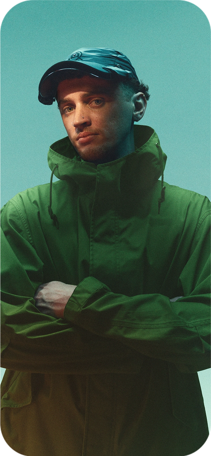Person wearing a dark green jacket and the Captured Cap, standing against a muted green background with arms crossed.