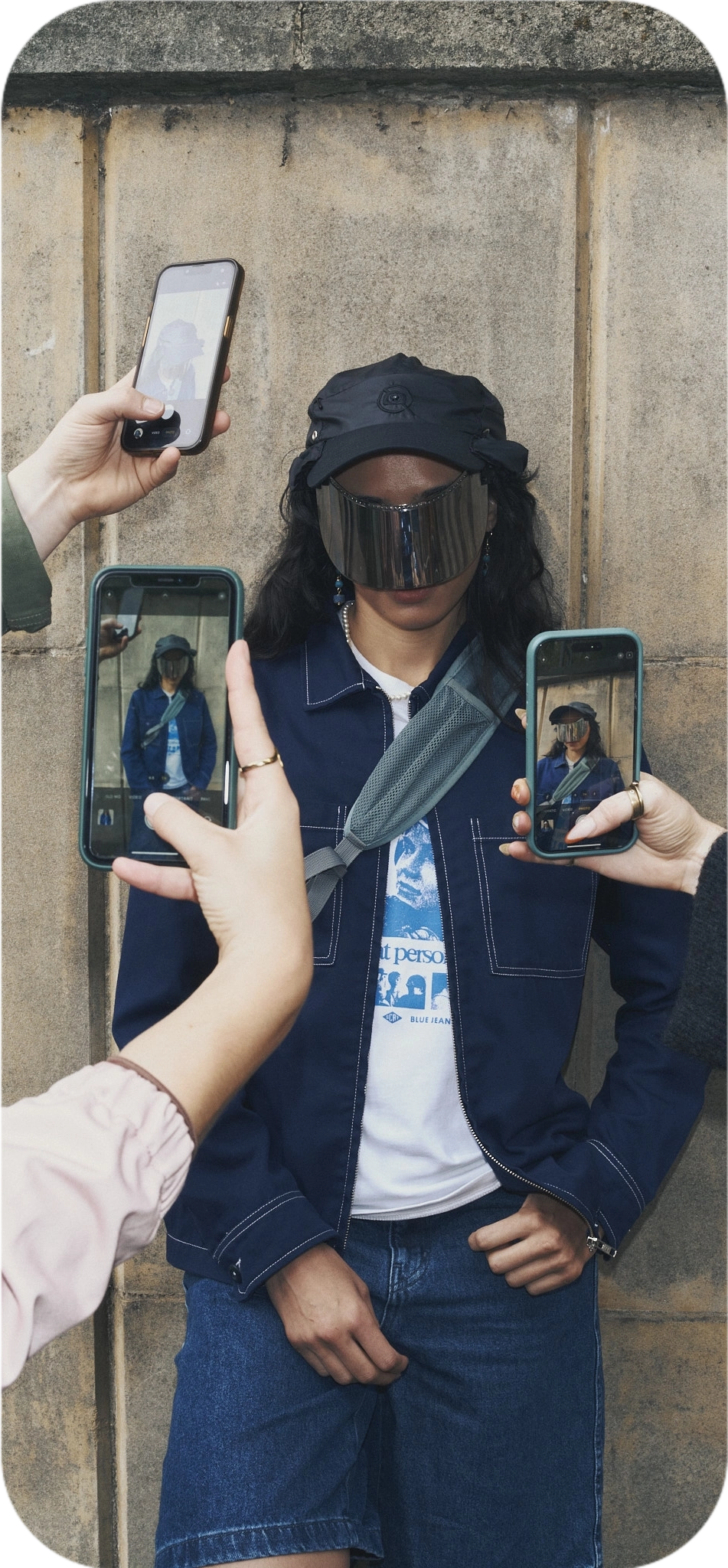 Person in a captured cap and dark jacket posing against a wall while several people photograph them with smartphones.